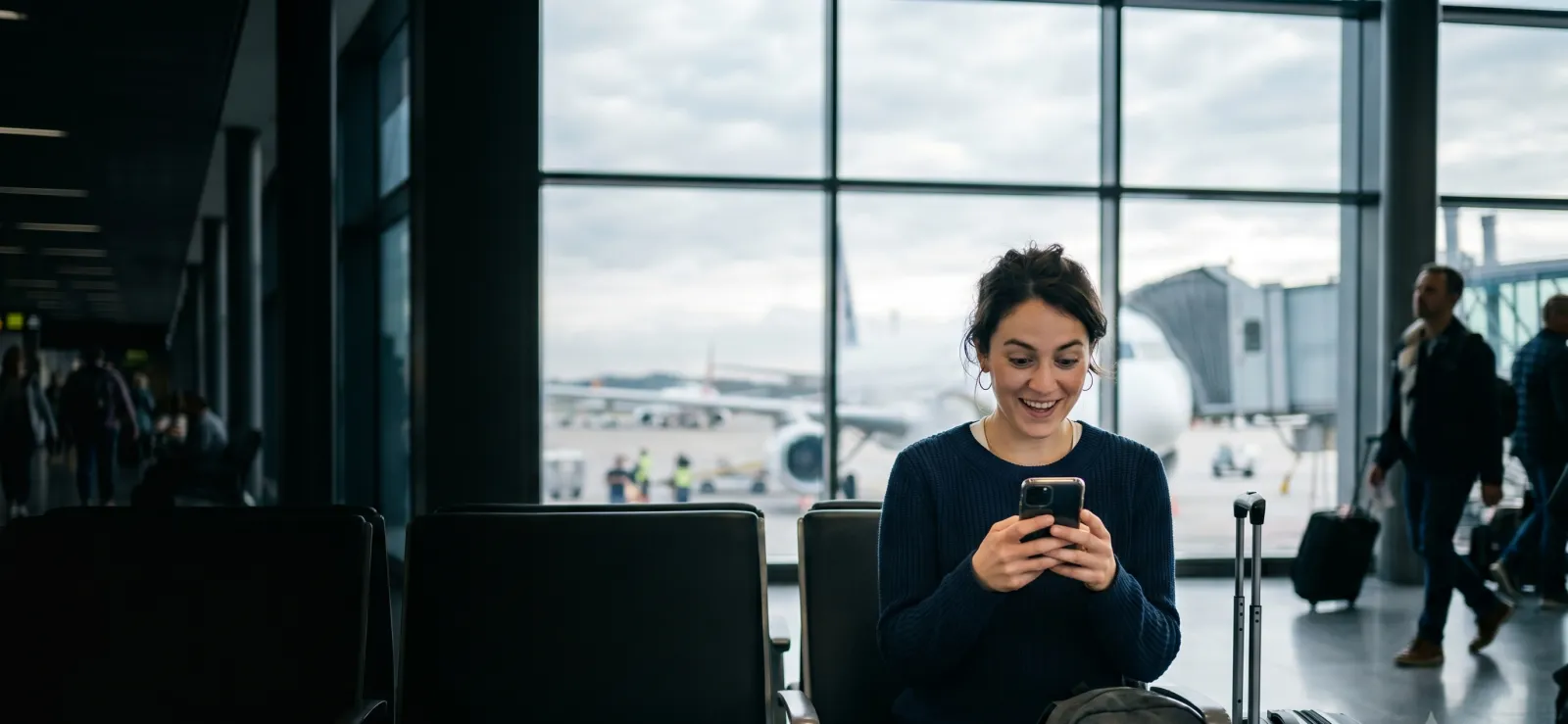 A traveler at an airport gate checking her phone, an aircraft at the window behind her